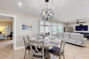 A modern dining area with a round table, chairs, and a chandelier. There is a sofa and a television in the background. - Canal-front home with screened pool, dock access, patio, grill, AC, & W/D (Cape Coral)