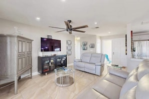A modern living room with a ceiling fan, a gray sofa, a glass coffee table, and a TV on a cabinet. - Canal-front home with screened pool, dock access, patio, grill, AC, & W/D (Cape Coral)