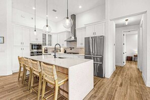 A modern kitchen with white cabinetry, a central island with bar stools, stainless steel appliances, and pendant lighting. - Newly-built luxury home with private infinity pool, canal views, & washer/dryer (Cape Coral)