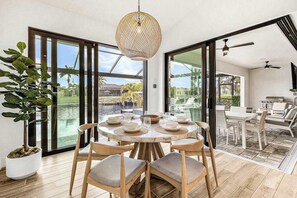 A dining area with a round table, chairs, a hanging light fixture, and large windows with a view of the outdoors. - Newly-built luxury home with private infinity pool, canal views, & washer/dryer (Cape Coral)