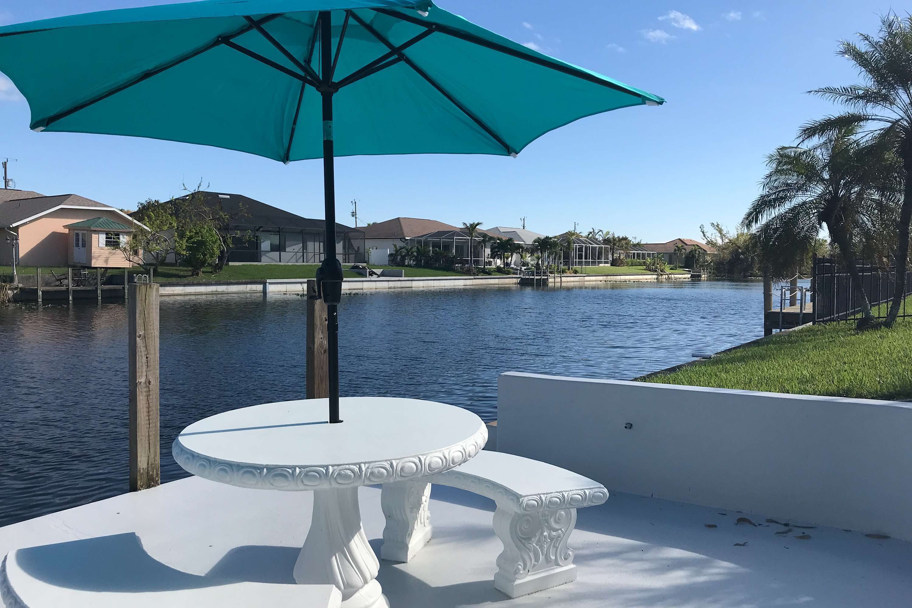 Une terrasse au bord de l’eau, avec une table et un banc blancs, un parasol turquoise et une vue sur les maisons et l’eau.