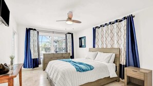 Un dormitorio con cama, mesita de noche, ventilador de techo y ventana con cortinas.