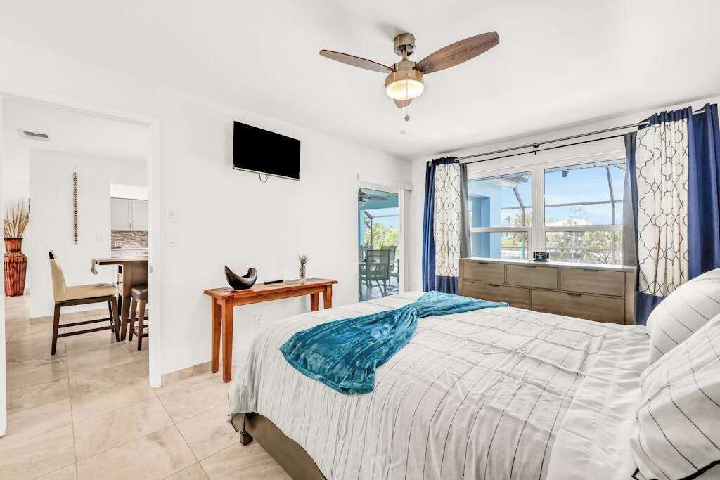 Une chambre à coucher comprenant un lit, un ventilateur de plafond, une télévision, une fenêtre avec des rideaux et un bureau avec une chaise.