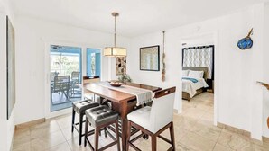 Un comedor con una mesa y sillas de madera, una lámpara colgante y vistas a un dormitorio con cama y cortinas.