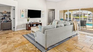 Ein Wohnzimmer mit grauem Sofa, einem Flachbildfernseher, einem Deckenventilator und Blick auf einen Pool durch Glastüren.