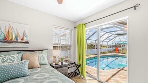 Ein Schlafzimmer mit Bett, Nachttisch, einem Gemälde, einem Fenster mit Jalousien und Blick auf einen Swimmingpool durch eine Glastür.