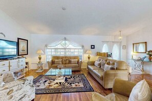 Ein geräumiges Wohnzimmer mit einer großen Couch, einem Couchtisch und einem Fernseher. Zwei Fenster mit Vorhängen, eine Kristallleuchter und ein dekorativer Spiegel sind ebenfalls vorhanden.