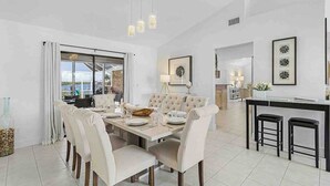 Uma sala de jantar ampla com uma mesa grande e cadeiras, área de cozinha com balcão, e vista através de uma porta de vidro.