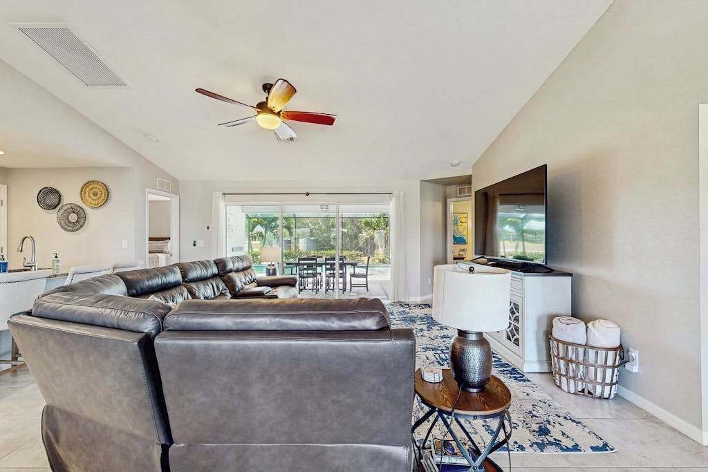 Un salon moderne avec un canapé d’angle, un ventilateur de plafond, un téléviseur et un coin cuisine visible à travers une porte ouverte.