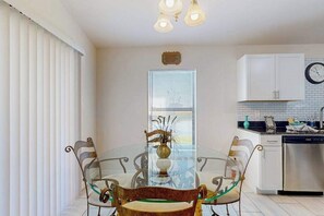 A dining area with a glass table, metal chairs, a vase, and a kitchen with cabinets and a dishwasher. - Cozy Home with backyard, canal view, fully equipped kitchen, dog friendly (Cape Coral)