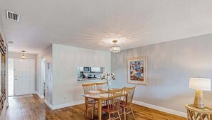 Un comedor amplio con sillas de madera y una mesa, una cocina al fondo y un cuadro en la pared.