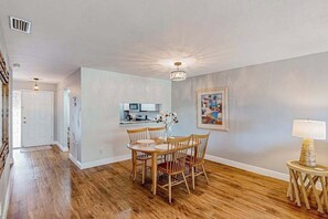 A spacious dining area with wooden chairs and a table, a kitchen in the background, and a painting on the wall. - Waterfront condo with balcony, pool, pickball/tennis, & walking paths (Fort Myers)