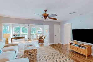 A spacious living room with a ceiling fan, a television, a wooden entertainment center, and a large window with a view of greenery. - Waterfront condo with balcony, pool, pickball/tennis, & walking paths (Fort Myers)