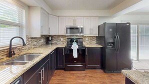 A modern kitchen with a granite countertop, stainless steel appliances, and dark cabinetry. - Bright and airy home with enclosed pool, hammock, and open-concept interior (Fort Myers)