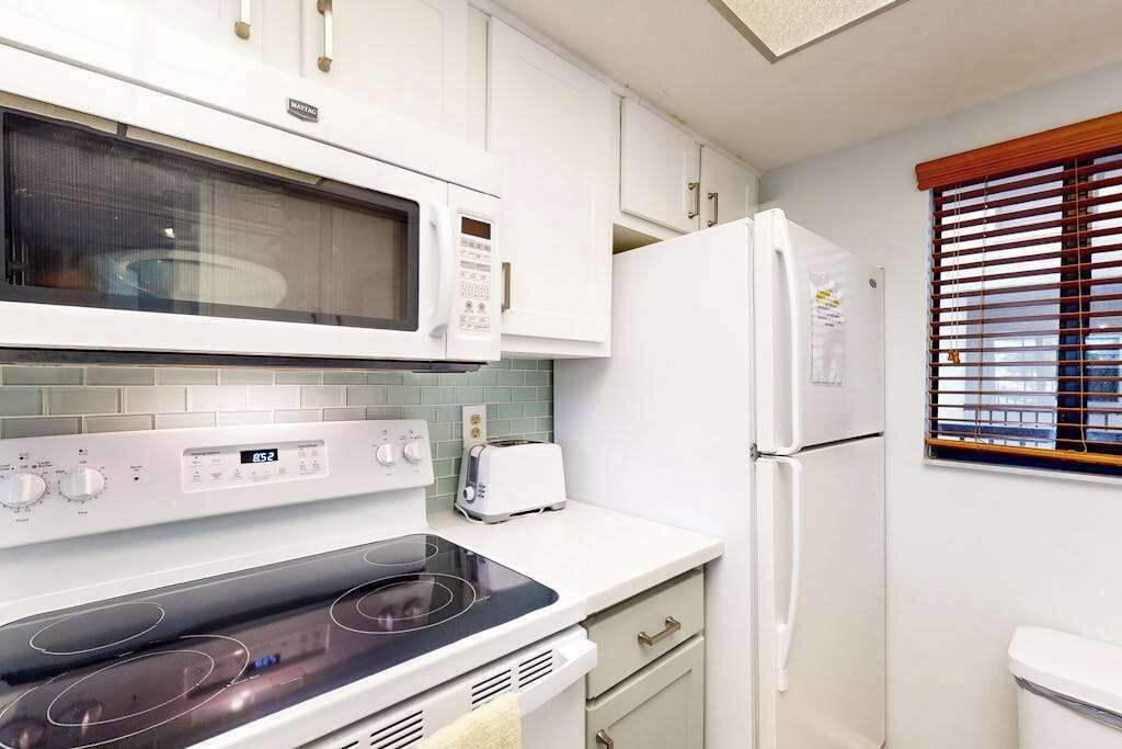 Een moderne keuken met een magnetron, fornuis, broodrooster, koelkast en een raam met jaloezieën.