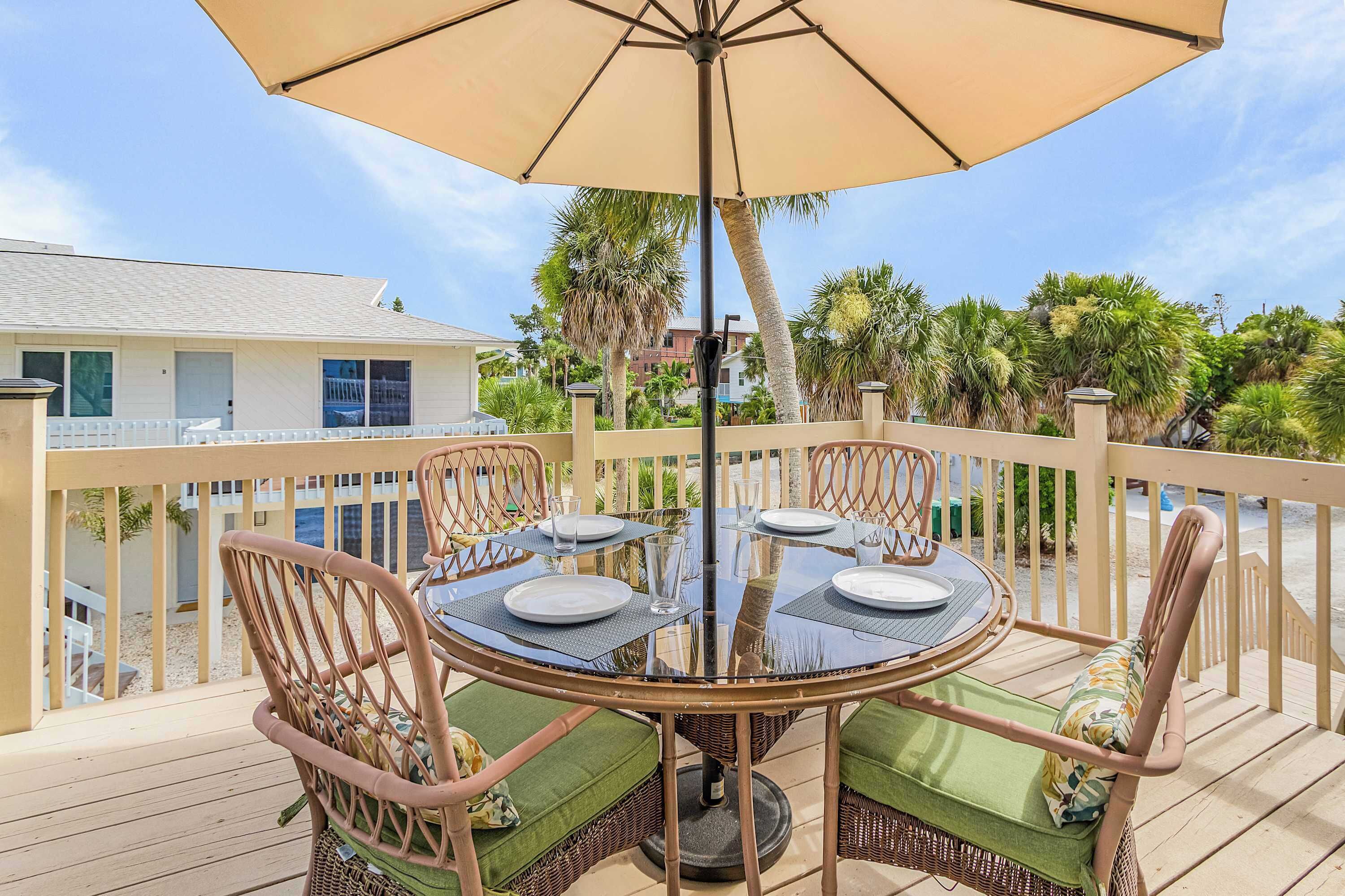 A deck with a round table and four chairs, an umbrella, and a view of a building and trees.