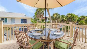 A deck with a round table and four chairs, an umbrella, and a view of a building and trees.