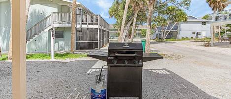 Una parrilla portátil con tanque de propano en un estacionamiento, con edificios al fondo.