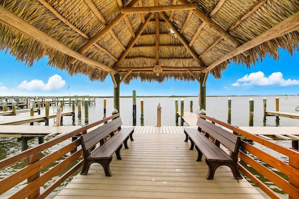 A wooden pier with benches, a thatched roof structure, and a view of the water.