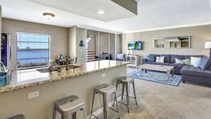 A modern living room with a kitchen area, a large window, and a flat-screen TV. - Amazing oceanfront condo with pool, hot tub, dock, & views - snowbird-friendly (Marco Island)