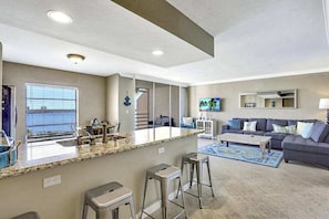 A modern living room with a kitchen area, a large window, and a flat-screen TV.