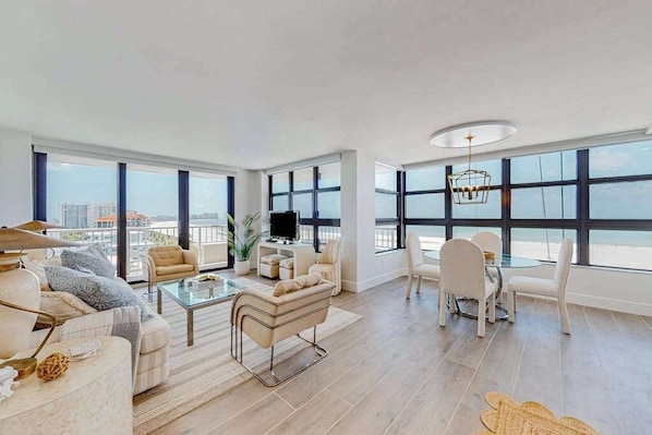 A spacious living room with a large window, a flat-screen TV, and a dining area with chairs and a table. - Gulf-front getaway with on-site pool, tennis, putting green, dock, & gym (Marco Island)