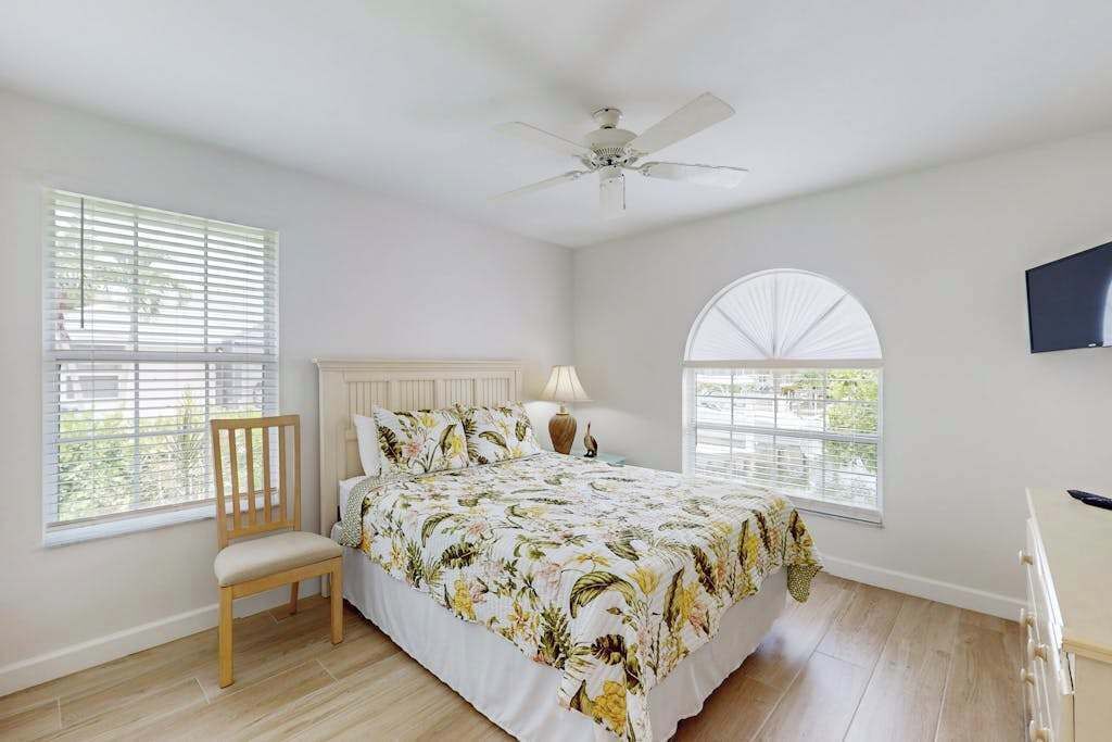 A bedroom with a bed, a chair, a ceiling fan, a TV, and a window with blinds.