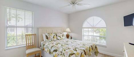 A bedroom with a bed, a chair, a ceiling fan, a TV, and a window with blinds.