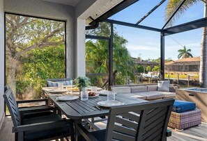 Un área de comedor al aire libre con una mesa y sillas, un espacio de descanso con cojines y vistas de la vegetación y edificios circundantes.