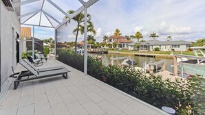 A covered outdoor area with a lounge chair, a view of a canal with boats, and houses in the background. - Waterfront home with private heated pool, spa, BBQ, views & bikes (Marco Island)