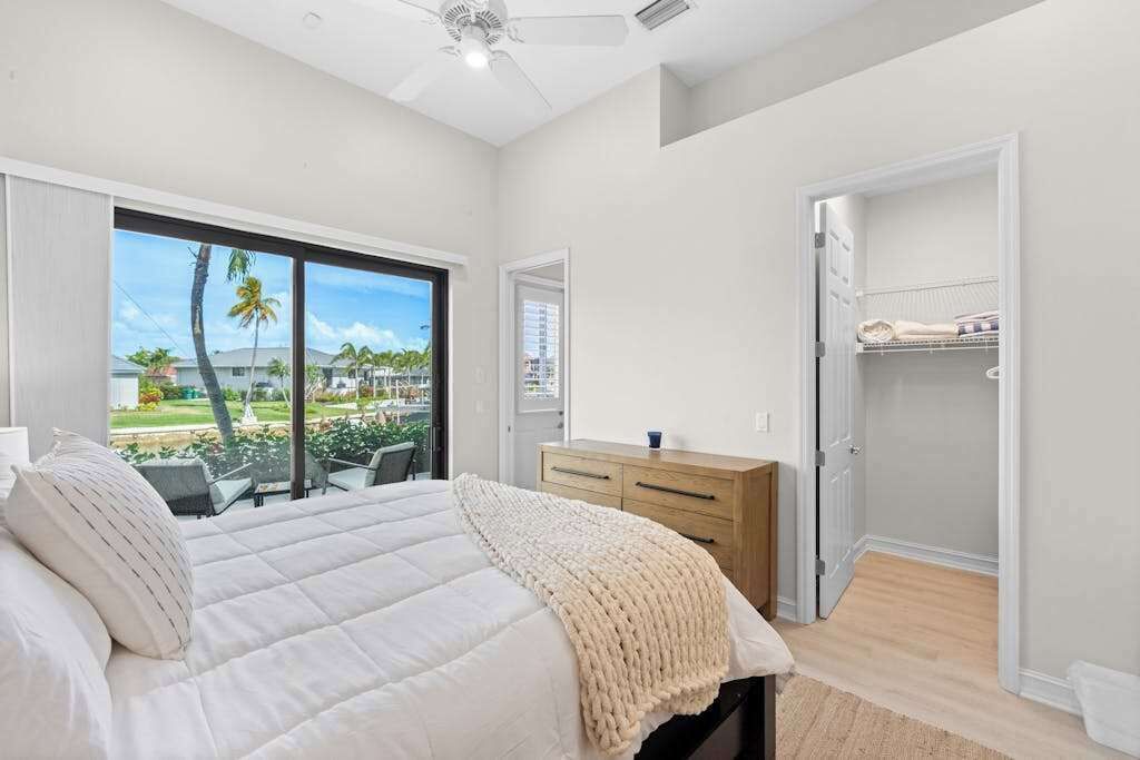 A bedroom with a bed, a wooden dresser, a closet, and a view of palm trees.
