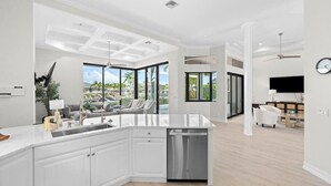 A modern kitchen with a central island, a dining area with a white sofa, and a view of a pool. - Waterfront home with private heated pool, spa, BBQ, views & bikes (Marco Island)