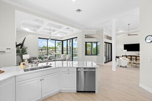 A modern kitchen with a central island, a dining area with a white sofa, and a view of a pool. - Waterfront home with private heated pool, spa, BBQ, views & bikes (Marco Island)