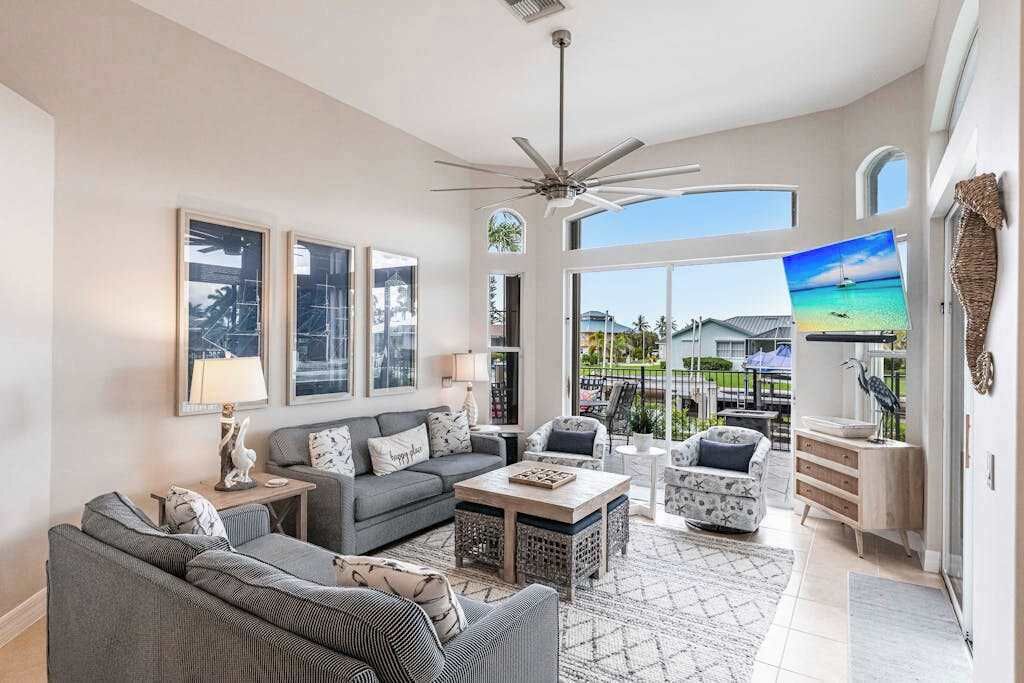 A spacious living room with a ceiling fan, large windows, a flat-screen TV, and a view of the outdoors.