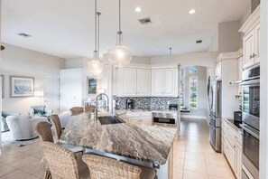 A modern kitchen with white cabinets, a marble island, stainless steel appliances, and wicker furniture. - Luxurious, waterfront home with private pool/spa (Marco Island)