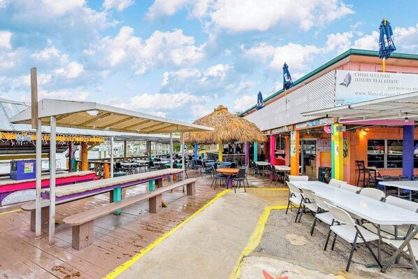 An outdoor dining area with tables and benches, a thatched-roof structure, and a building with a sign for a real estate business.