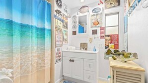 A bathroom with a beach-themed shower curtain, a white vanity with a mirror, and various wall decorations including a fish and a poster. - Waterfront home with kayaks, washer/dryer & fast WiFi (Goodland)