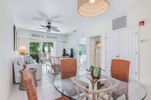 A modern interior with a glass dining table, wicker chairs, a ceiling fan, and a sign reading 'MARCO ISLAND'. - Renovated condo with screened balcony, AC, & shared pool/spa/sundeck (Marco Island)