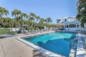 A swimming pool with lounge chairs, palm trees, and a building with a white facade. - Renovated condo with screened balcony, AC, & shared pool/spa/sundeck (Marco Island)