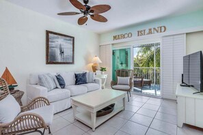 A living room with a white sofa, wicker chairs, a coffee table, a ceiling fan, a framed picture, and a TV on a cabinet. The room has a balcony with a view of palm trees. - Renovated condo with screened balcony, AC, & shared pool/spa/sundeck (Marco Island)