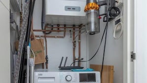A compact laundry room with a front-loading washing machine, a water heater, and various pipes and hoses. - New 1 Bed Flat Near Central London With Terrace (London)