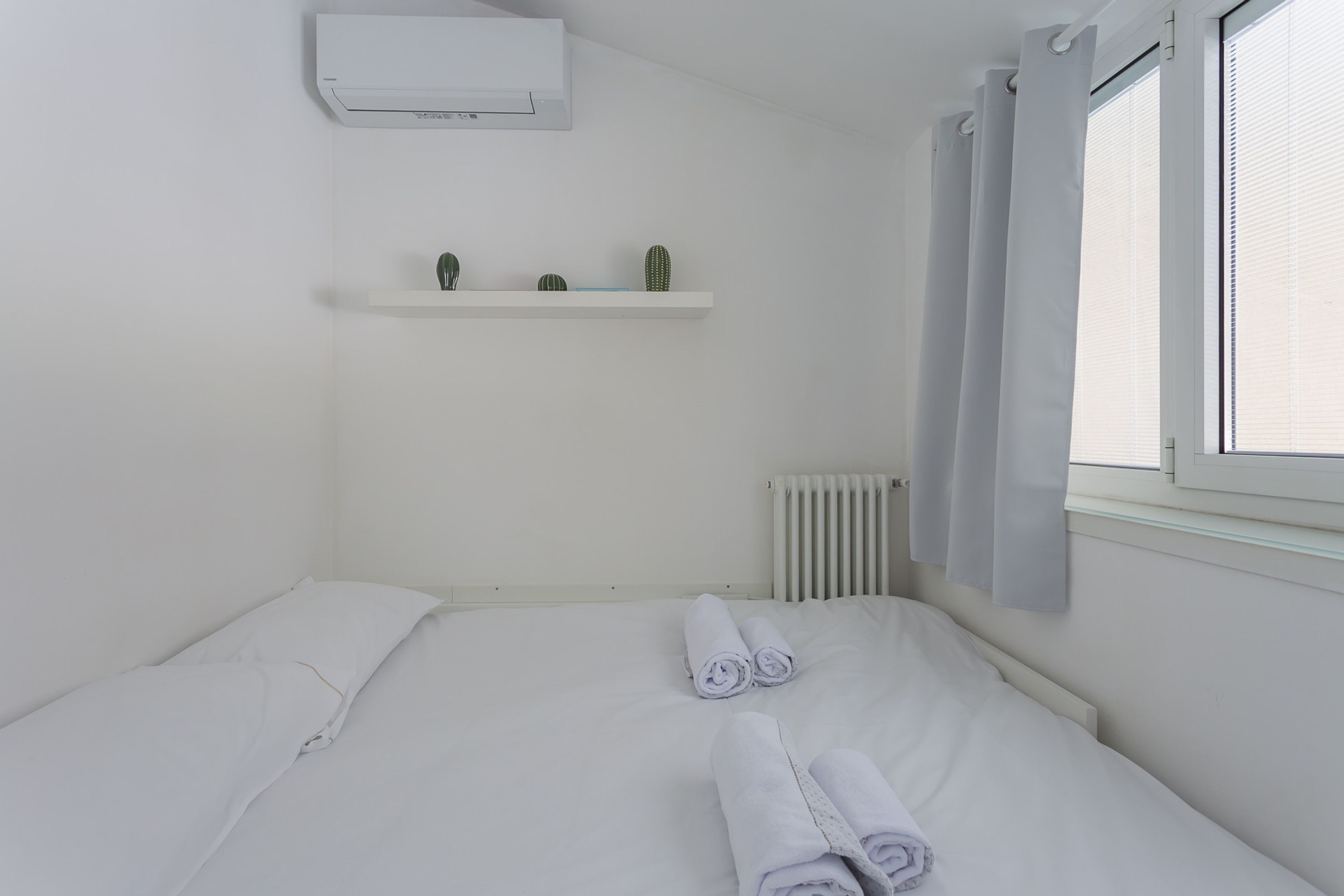 Una cama individual con toallas enrolladas, un aire acondicionado blanco, un radiador y una ventana con cortinas.
