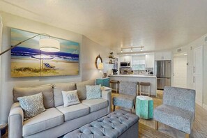Ein Wohnzimmer mit einer Couch, Sesseln, einem Couchtisch, einem Gemälde und einer Küche im Hintergrund.
