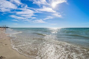 Uma praia com céu claro, águas calmas e um píer ao longe.