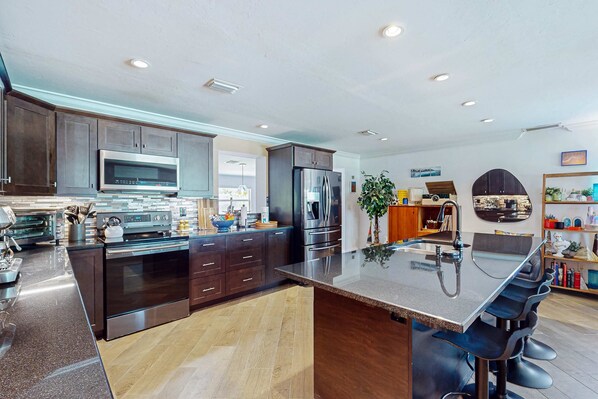 A modern kitchen with dark cabinetry, stainless steel appliances, and a central island with bar stools. - Updated home near beach/golf with private yard, firepit, & home office (Naples)
