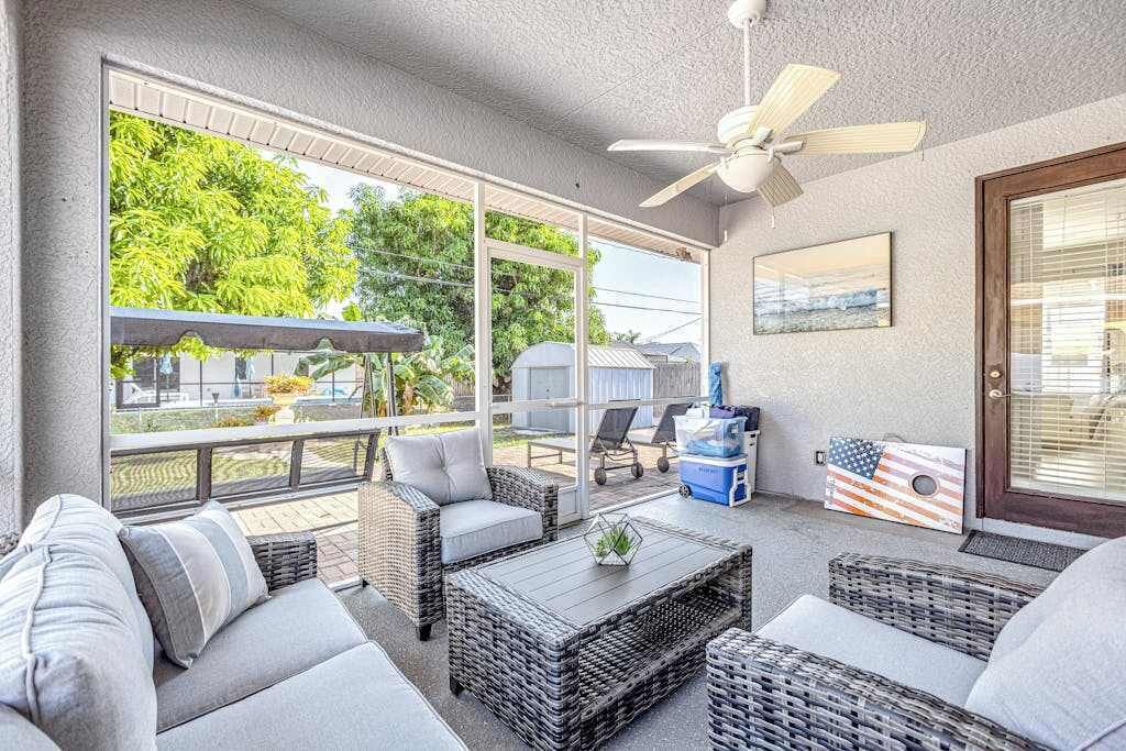 En overdækket terrasse med fletningsmøbler, ventilator i loftet og store vinduer der giver udsigt til grønne områder.