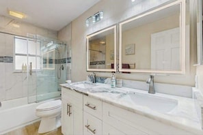 Une salle de bain dotée d’une douche à parois de verre, de toilettes blanches, d’un lavabo double avec un plan de travail en marbre et d’un grand miroir.