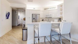 A modern kitchen with white cabinetry, a stainless steel refrigerator, a microwave, and a sink. There are three white bar stools and a trash can. - Luxurious condo with on-site pool, private extended balcony, & W/D (Naples)
