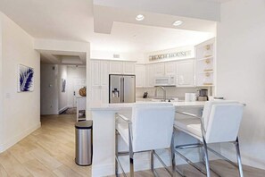 A modern kitchen with white cabinetry, a stainless steel refrigerator, a microwave, and a sink. There are three white bar stools and a trash can. - Luxurious condo with on-site pool, private extended balcony, & W/D (Naples)