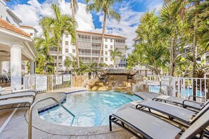 A hotel pool area with lounge chairs, palm trees, and a rock waterfall feature. - Luxurious condo with on-site pool, private extended balcony, & W/D (Naples)